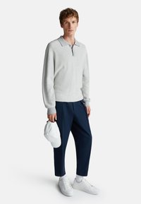 Lichtgrijze, lange mouwen polo met een kraag, marineblauwe trousers en witte sneakers. Witte pet in de hand gehouden, met een zichtbaar, gestructureerd stofdetail.