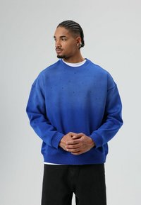 Sweat-shirt bleu avec un design dégradé, orné de petits clous noirs, col rond et poignets côtelés. Porté par-dessus une chemise blanche.