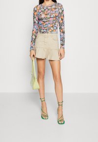 Långärmad blommig topp i livliga färger, beige rynkad minikjol, gröna sandaler med remmar och en liten ljusgrön väska.