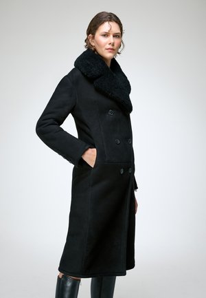 Femme portant un long manteau noir à double boutonnage avec un large col en shearling et des bottes noires, debout avec une main dans la poche.