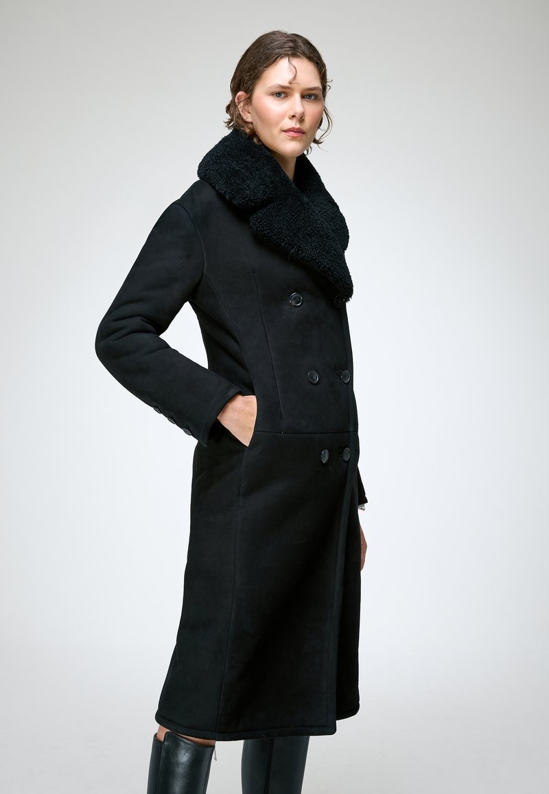Femme portant un long manteau noir à double boutonnage avec un large col en shearling et des bottes noires, debout avec une main dans la poche.