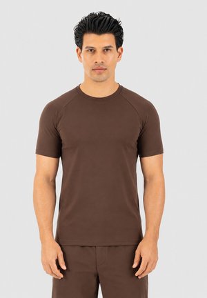 Hombre con pelo oscuro, vestido con una camisa marrón ajustada de manga corta y pantalones a juego, de pie frente a un fondo liso gris claro.