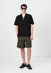 Cămașă neagră cu mâneci scurte și guler cu fermoar, asortată cu pantaloni scurți de culoare verde măsliniu și pantofi din piele negri. Ochelarii de soare completează ținuta.