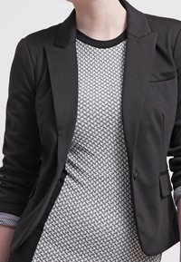Blazer nero con dettagli sui rever, chiusura a un bottone e due tasche frontali. Sotto, un top a maniche corte a righe grigie e bianche con collo girocollo.