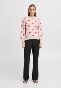 Pull rose clair avec des motifs de cœurs roses foncés éparpillés, col rond et manches bouffantes, associé à un pantalon noir à jambe droite.
