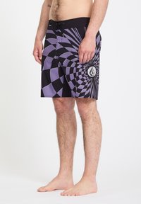 Homme portant un short de bain à motifs géométriques noirs et violets, debout pieds nus devant un fond blanc.