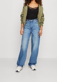 Oliwkowo-zielona kurtka bomberka na czarnym topie na ramiączkach z koronkowym wykończeniem, w zestawieniu z jasnoniebieskimi, szerokimi spodniami dżinsowymi i białymi trampkami z beżowymi akcentami.