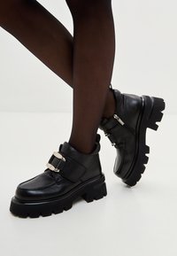 Botines de cuero negro con suela gruesa, con una correa ancha con hebillas plateadas y cremallera lateral. Textura lisa y diseño resistente.