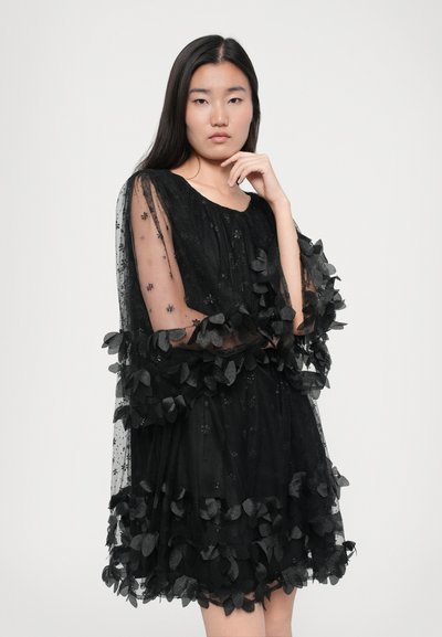 Zwarte jurk met doorschijnende mouwen, versierd met stoffen bloemen en kantdetails. Uitlopende silhouet, gestructureerde stof en opvallende vlinderaccenten.