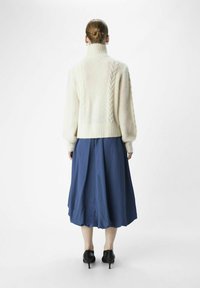 Cremfarbener Strickpullover mit hohem Kragen, kombiniert mit einem knielangen blauen Rock. Schwarze hohe Schuhe. Texturiertes Kleidungsstück und lockere Passform.