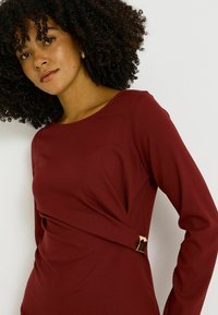 Robe à manches longues bordeaux avec un design drapé et un accent en métal doré à la taille, fabriquée dans un tissu lisse et extensible.