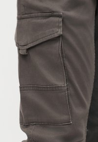 Tamsiai pilkos audinio kargo kelnės kišenė su atvartėliu ir matomomis siūlių detalėmis ant kojos šono.