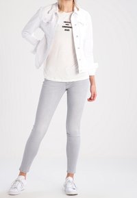 Chaqueta de mezclilla blanca sobre una camiseta gráfica clara con texto. Unos jeans skiny gris claro y unas zapatillas blancas completan el atuendo.