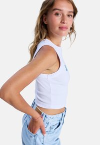 Witte gecropte tanktop met ribbeltextuur, mouwloos ontwerp en blauwe bloemenborduring. Draag met lichtblauwe high-waisted denim shorts.