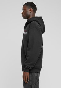 Sudadera negra con bolsa canguro en el frente, capucha con cordón, puños acanalados y detalle de logo bordado. Modelo que lleva pantalones gris oscuro.