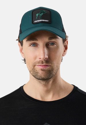 Man met lichte baard, draagt een donker groene en zwarte pet met elandlogo en een zwart shirt, tegen een effen lichte achtergrond.