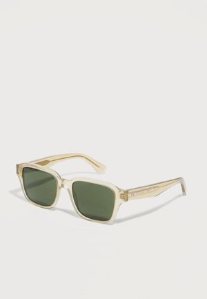 Lunettes de soleil rectangulaires transparentes beige avec des verres vert foncé reposant sur une surface blanche.