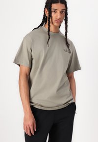 Camiseta verde oliva de manga corta hecha de algodón, con un pequeño diseño gráfico negro en la parte delantera, corte relajado y cuello redondo.