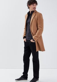 Kamelhaar-Mischmantel über einem grauen Pullover, schwarzen Hosen und schwarzen Sneakers. Der Mantel hat einen Ein-Knopf-Verschluss und einen Reverskragen.