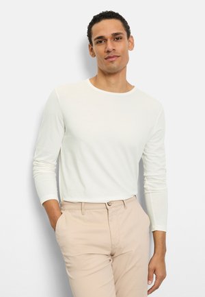 Camicia bianca a maniche lunghe in tessuto morbido, con scollo arrotondato. Abbinata a pantaloni beige chiaro, caratterizzati da una vestibilità comoda.
