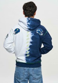 Huvtröja med en tie-dye design i blått och vitt, med amerikansk fotbollshjälm grafik och "COVERED" text i grönt på baksidan.