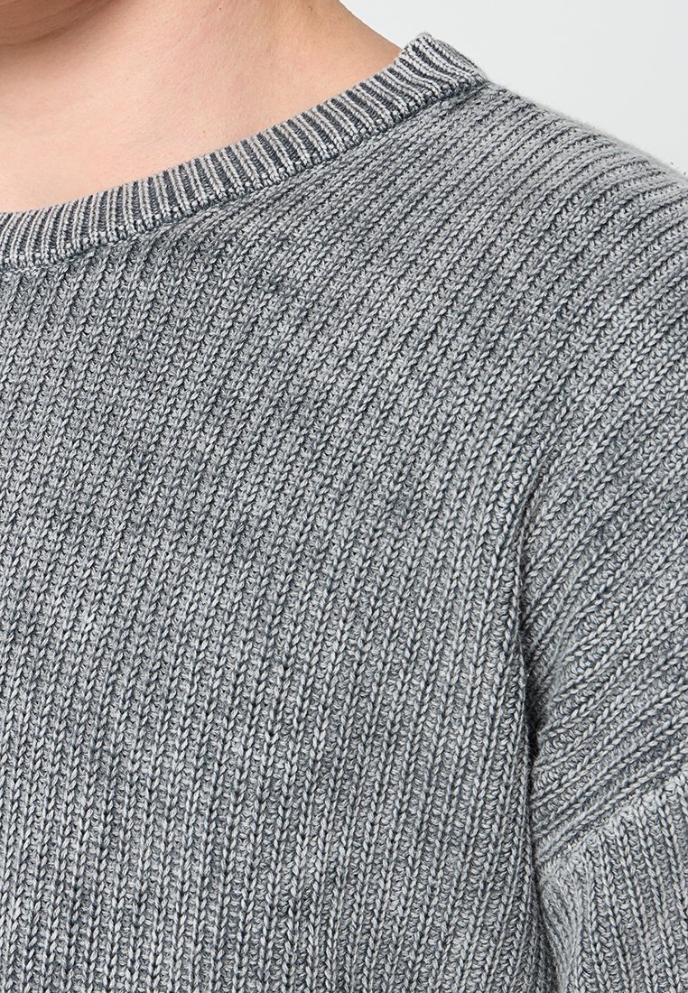 Close-up van een persoon die een grijze geribbelde gebreide trui met een ronde halslijn draagt.