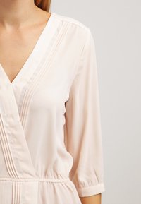 Jasnoróżowa bluzka zapinana z dekoltem w serek, trzy czwarte rękawów, pionowymi plisami i elastyczną talią dla dopasowanego sylwetki.