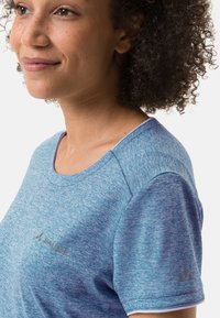 Camisa de manga corta azul mélange con escote redondo, que presenta un logo metálico en el pecho y un acento en forma de triángulo en la manga.