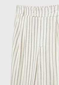 Pantalon en lin beige avec fines rayures noires verticales, taille haute, plis, poches latérales et deux boutons beiges à la taille.
