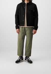Sort, knaplukket jakke med lange ærmer og lommer foran. Beige crewneck t-shirt under. Olivengrønne, løse bukser i struktureret stof. Sorte sneakers.