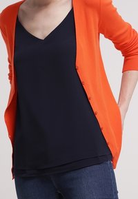 Cardigan arancione con chiusura a bottoni sopra un top blu navy a scollatura a V. Entrambi i capi sono realizzati in un tessuto morbido, con una vestibilità rilassata e una consistenza liscia.