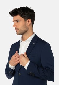 Marineblauer Anzugsakko mit Reverskragen, Ein-Knopf-Verschluss vorne und feiner Struktur. Das Modell trägt darunter ein weißes Hemd mit sichtbaren Manschetten.