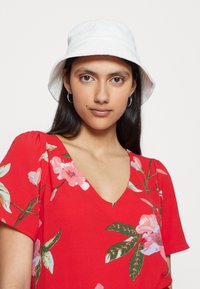 Vestido vermelho com padrão floral e mangas curtas bufantes, apresentando flores rosas e verdes. Chapéu estilo bucket branco, de textura suave, usado para proteção solar.