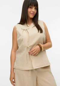 Beige mouwloos topje met een V-hals en striksluitingen aan de voorkant, gemaakt van een gestructureerde stof. Gecombineerd met bijpassende losse broek.