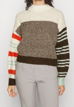 Frau trägt einen mehrfarbigen Strickpullover mit gemischten braunen, cremefarbenen, orangefarbenen und minzfarbenen Streifen, kombiniert mit hellgrauen Hosen.