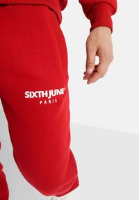 Rode sweatpants gemaakt van zachte stof, met een witte geborduurde "SIXTH JUNE PARIS" logo op de linkerdij, zijzak zichtbaar.