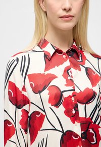 Knoopshirt van lichtgewicht stof, met een witte basis en grote rode bloempatronen met zwarte omlijningen, lange mouwen en een kraag.