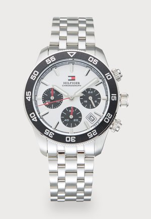 Montre chronographe en acier inoxydable avec cadran blanc, sous-cadrans noirs, lunette noire et bracelet argenté. Présente des accents rouges et un affichage de la date.