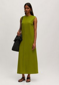 Langes, ärmelloses Kleid aus lebhaftem grünen Stoff, mit geradem Schnitt und rundem Ausschnitt. Ergänzt durch eine schwarze, geflochtene Tasche mit Fransen.