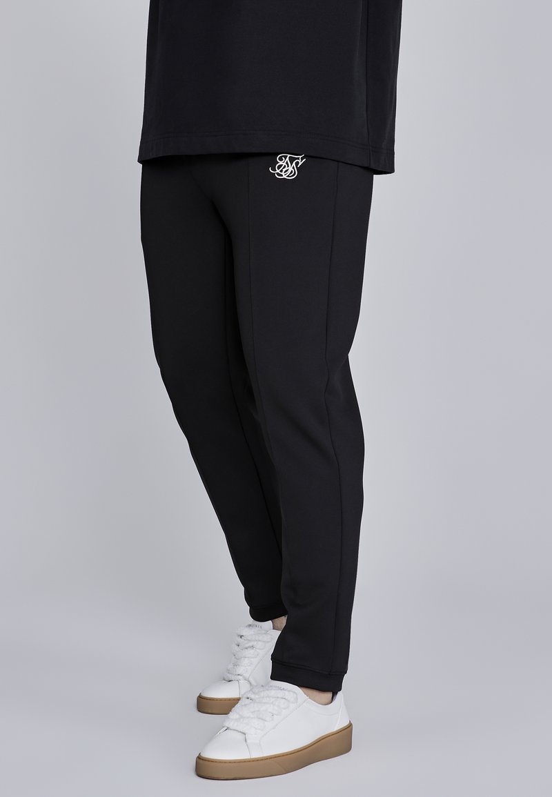 Personne portant un jogging noir slim avec un logo blanc sur la cuisse et des baskets blanches avec des semelles en gomme, debout sur un fond gris.