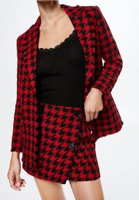 Conjunto de blazer y falda con patrón de pata de gallo en rojo y negro. El blazer tiene un solo botón y la falda presenta dos botones negros. Debajo, una camiseta sin mangas negra.