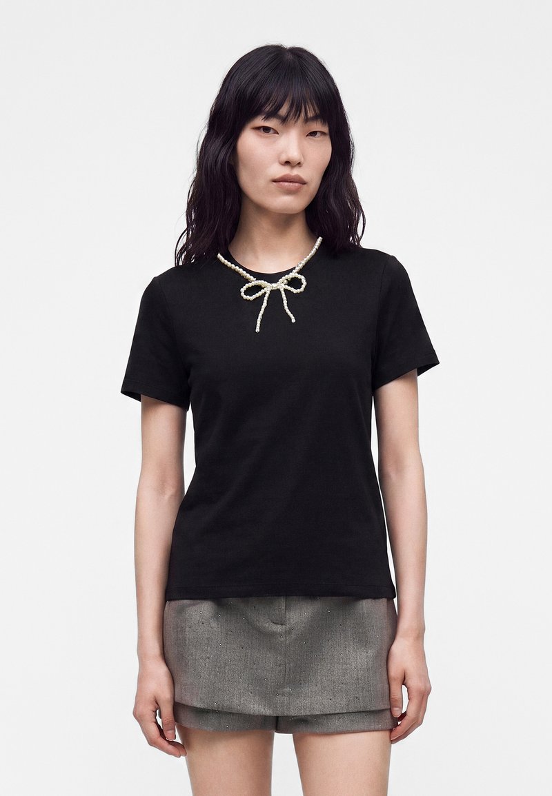 Femme aux cheveux noirs portant un haut noir à manches courtes, un collier en forme de nœud de perles et un short gris taille haute.