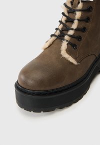 Bota de cuero marrón hasta el tobillo con cordones negros, forro de piel sintética y una suela de goma negra texturizada con un diseño de banda elevada.
