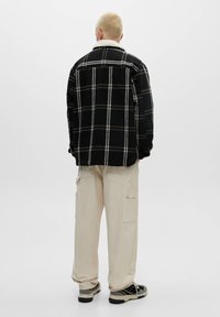 Persona con capelli biondi corti che indossa una giacca a scacchi bianca e nera, pantaloni cargo beige e sneakers nere, in piedi con le spalle rivolte.