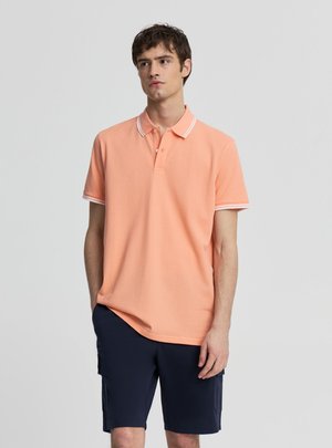 Młody mężczyzna w brzoskwiniowej koszulce polo z krótkim rękawem z białą lamówką i granatowych spodenkach typu cargo, stojący z jedną ręką w kieszeni.