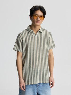 Giovane uomo che indossa occhiali con lenti arancioni, una polo a maniche corte a righe lavorata a maglia e jeans azzurri, in piedi davanti a uno sfondo semplice.