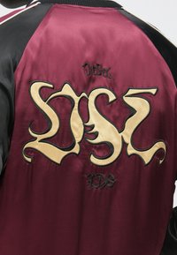 Burgundia satiinist bomberjakk mustade varrukatega, millel on suured kreemjad tähed ja seljal tikitud "Diesel" logo.