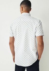 Camisa de manga corta de tela blanca con estampado tropical en azul. Presenta un cuello estándar y dobladillo curvado.