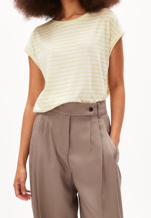 Persoon die een geel-wit gestreept shirt met korte mouwen draagt, ingestopt in een losse, hooggetailleerde taupekleurige broek met plooien en een tailleband met knopen.