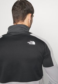 Giacca zip-up nera e grigia con colletto alto, dotata di un tessuto testurizzato e di un logo bianco sulla parte superiore della schiena.